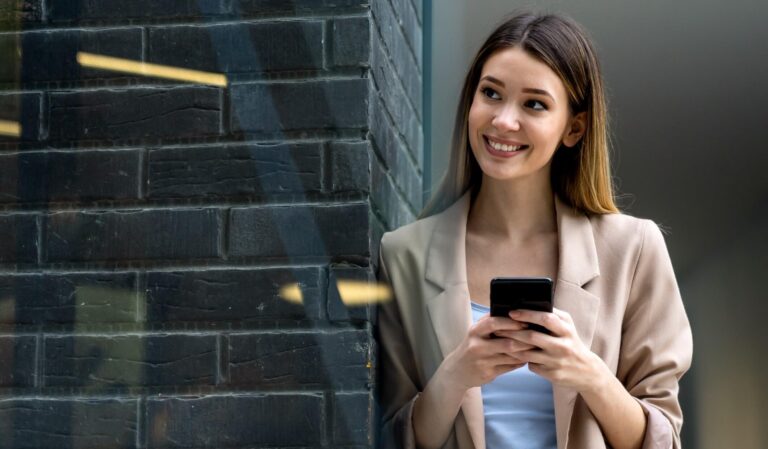 Instagram Strategie, Frau mit Smartphone in der Hand steht angelehnt an eine dunkle Backsteinmauer